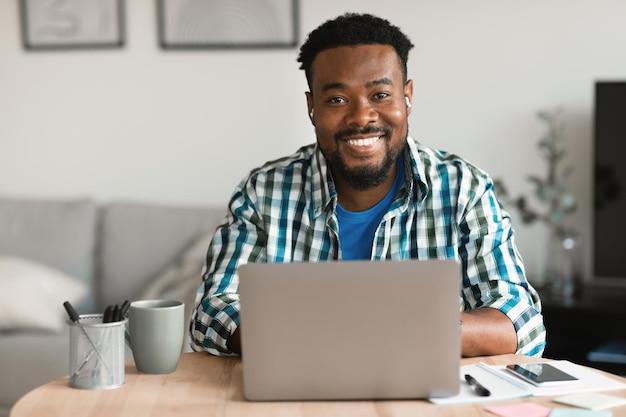 Homem negro alegre no laptop usando fones de ouvido trabalhando dentro de casa