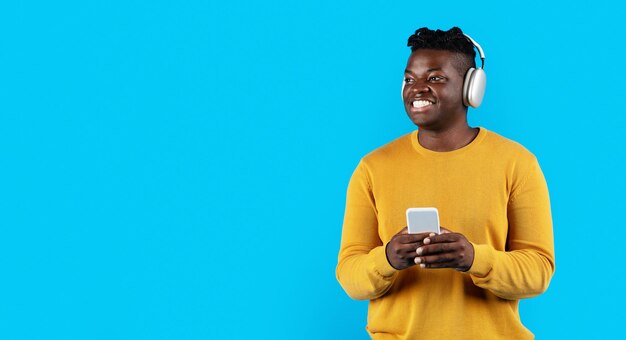 Homem negro alegre usando fones de ouvido segurando smartphone e olhando de lado