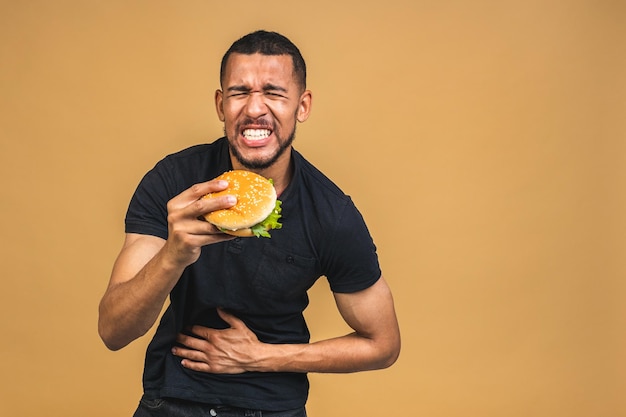 Homem negro americano africano faminto comendo hambúrguer isolado sobre fundo verde dor de estômago