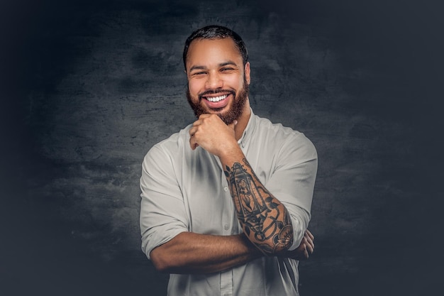 Homem negro barbudo positivo com uma tatuagem nos braços cruzados, vestido com uma camisa branca.