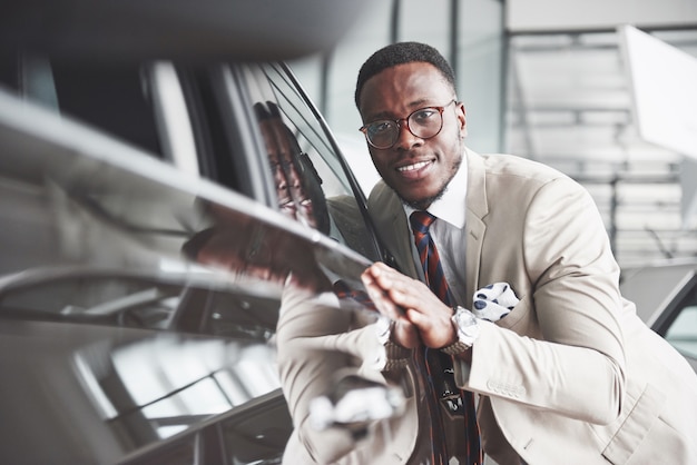 Homem negro bonito na concessionária está abraçando seu carro novo e sorrindo