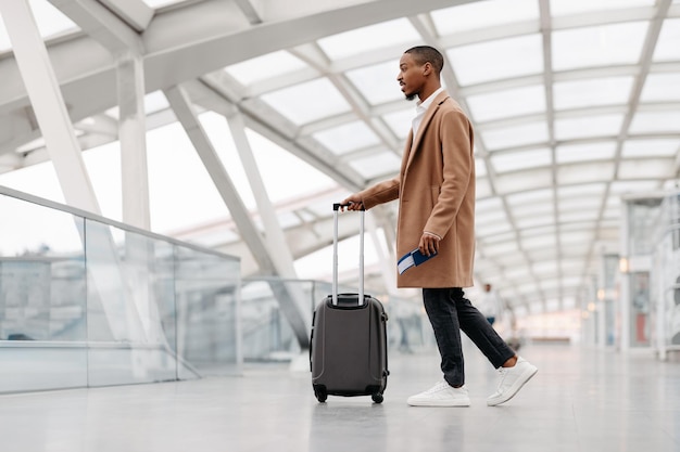 Homem negro bonito vestindo casaco elegante andando com mala no aeroporto