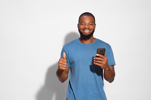 Homem negro com uma camiseta azul e um telefone em uma mão sorrindo e mostrando como gesto