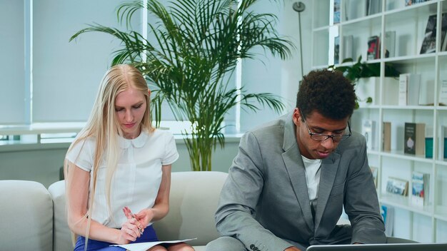 Homem negro compartilhando boas notícias com colegas de trabalho. Homem afro-americano surfando no laptop e compartilhando ótimas notícias com colegas de trabalho sentados juntos no escritório ao redor da mesa.