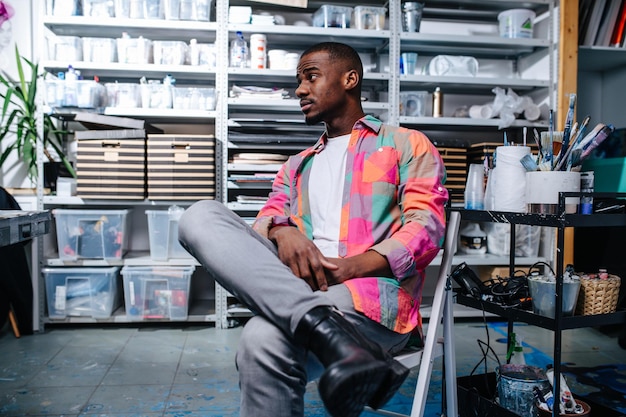 Homem negro descontraído em uma camisa estampada quadrada colorida, sentado em uma cadeira em uma oficina. Todos os tipos de recipientes nas prateleiras no fundo.