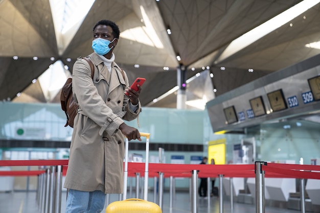 Homem negro em pé no aeroporto usando máscara protetora durante a epidemia de vírus, covid-19