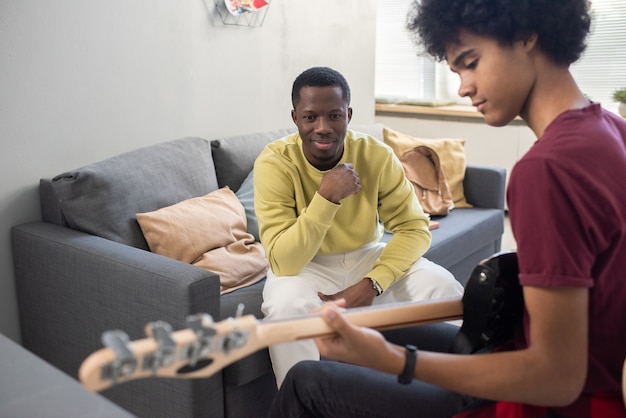 Homem negro feliz em trajes casuais olhando para um rapaz tocando violão durante a aula de música em casa