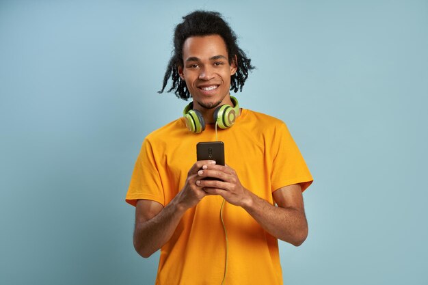 Homem negro navega na internet no celular em fundo azul, vestido casualmente