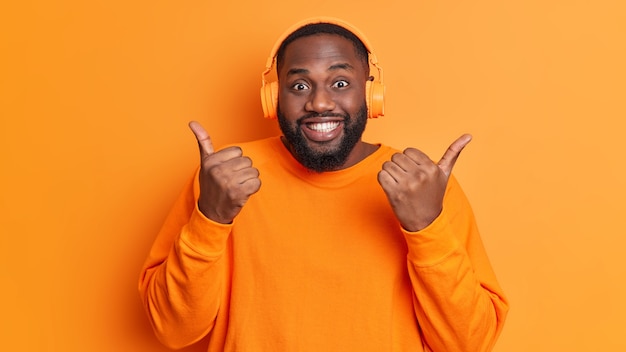 Homem negro positivo com barba densa faz gesto de gesticular para cima Desfruta da lista de faixas favoritas em fones de ouvido e usa um macacão casual de manga comprida isolado sobre uma parede laranja viva