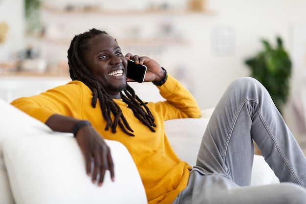 Homem negro positivo falando no celular enquanto relaxa no sofá em casa
