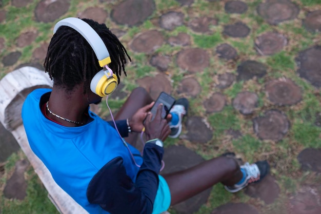 homem negro sentado em um playground em roupas esportivas, ouvindo música com fones de ouvido pelo telefone