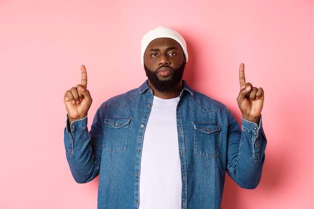 Homem negro sério e incomodado com barba, olhando para a câmera e apontando os dedos para cima, mostrando a promo, em pé sobre um fundo rosa.