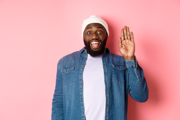 Homem negro sorridente e amigável dizendo olá, acenando com a mão, cumprimentando você, de pé sobre um fundo rosa