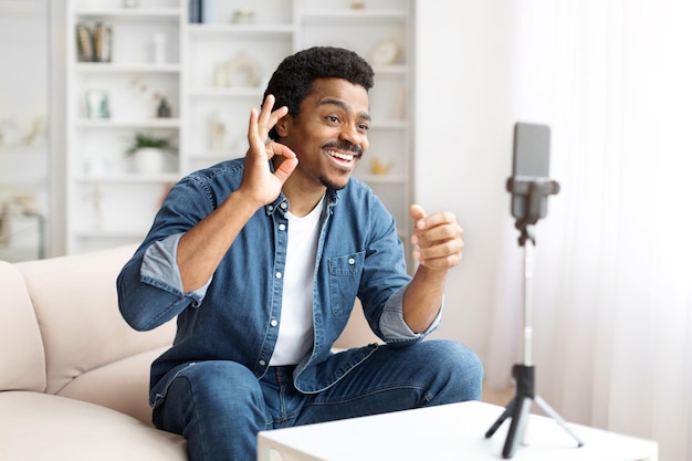 Homem negro sorridente fazendo um gesto ok durante uma videochamada em casa