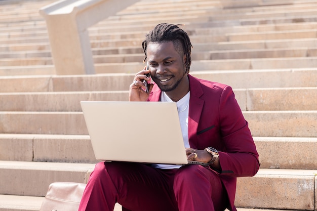 Homem negro sorridente trabalhando com computador, laptop e telefone sentado nas escadas da cidade
