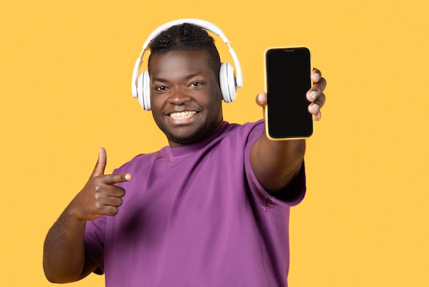 Homem negro usando fones de ouvido mostrando smartphone e como fundo amarelo