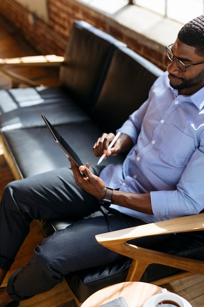 Foto homem negro usando uma caneta com um tablet digital em um modelo social de sofá