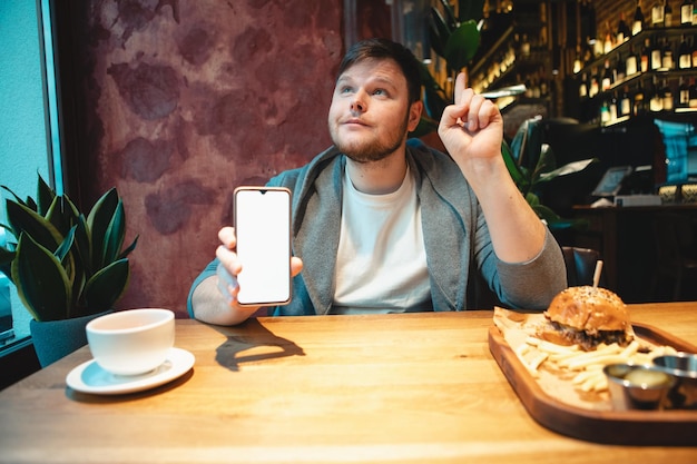 Homem no café segurando o telefone com tela branca comendo hambúrguer