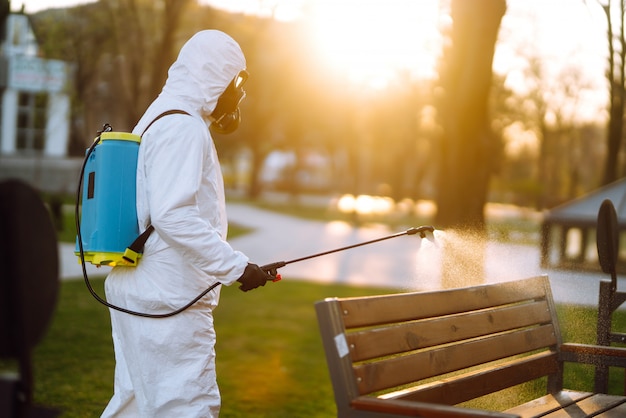 Homem no terno protetor que desinfeta o banco no parque público na cidade de quarentena. Prevenção de infecções e controle de epidemias. Covid 19.