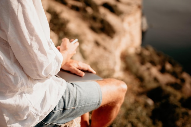 Homem nômade digital de chapéu um empresário com um laptop sentado nas rochas à beira-mar durante o pôr do sol