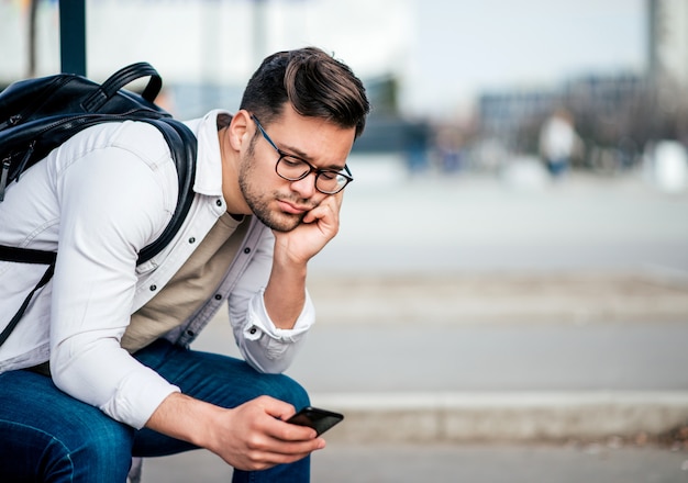 Homem novo entediado ocasional com o backcpack que senta-se no banco e que espera o ônibus, usando o smartphone.