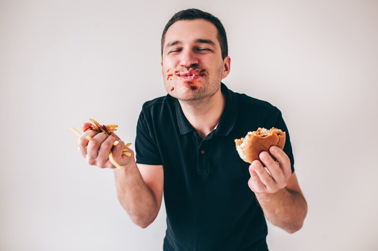 Foto homem novo isolado sobre a parede branca. cara feliz satisfeita com ketchup ao redor da boca sorrindo. segure o hambúrguer e as batatas fritas nas mãos.