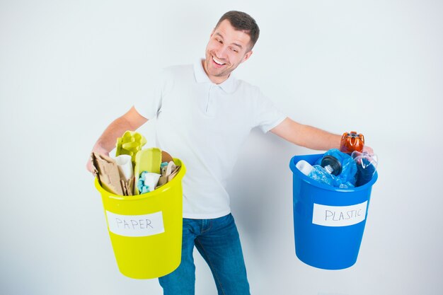 Homem novo isolado sobre a parede branca. Cara segura baldes coloridos pesados com plástico classificado e resíduos de papel. Tome cuidado com o meio ambiente. Preparação de materiais para reciclagem.