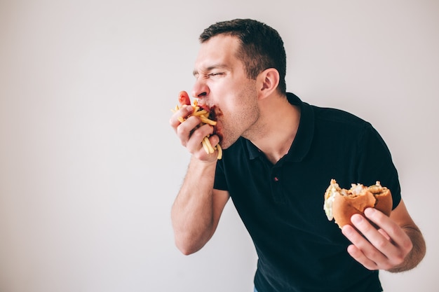 Homem novo isolado sobre a parede branca. Glutton cara devorando batatas fritas, que é realizada na mão. Hambúrguer mordido em outro. Gostoso delicioso lanche ou refeição comendo.