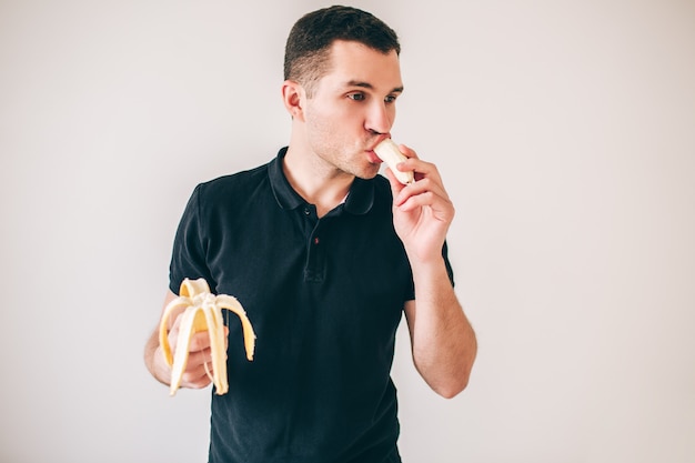 Homem novo isolado sobre o branco. Cara comendo banana com prazer. Segure duas peças separadas em mãos diferentes.