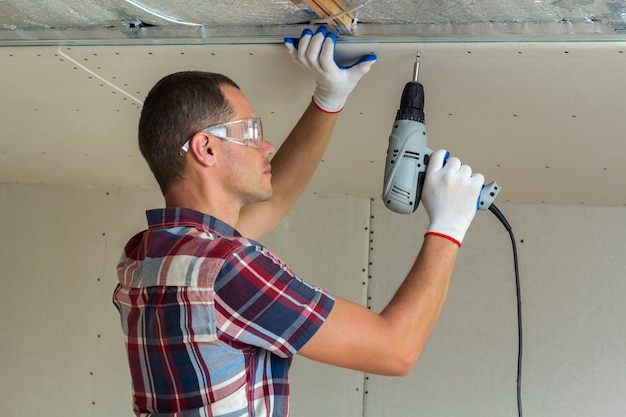 Homem novo nos óculos de proteção que fixa o drywall suspendeu o teto à armação de metal usando a chave de fenda elétrica no teto isolado com folha de alumínio brilhante. Renovação, construção, faça você mesmo conceito.