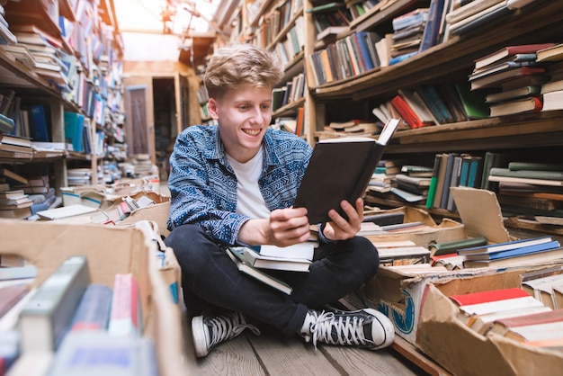 Homem novo positivo que senta-se no assoalho em uma biblioteca pública acolhedora, lendo livros e sorrindo.