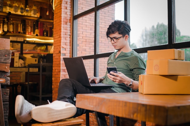 Homem novo que senta-se com computador e telefone celular no assoalho de madeira com o pacote que vende o conceito das ideias em linha.