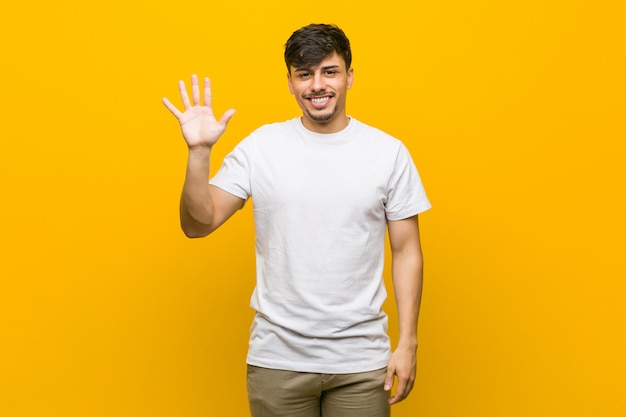 Homem ocasional latino-americano novo que sorri mostrando alegre número cinco com dedos.