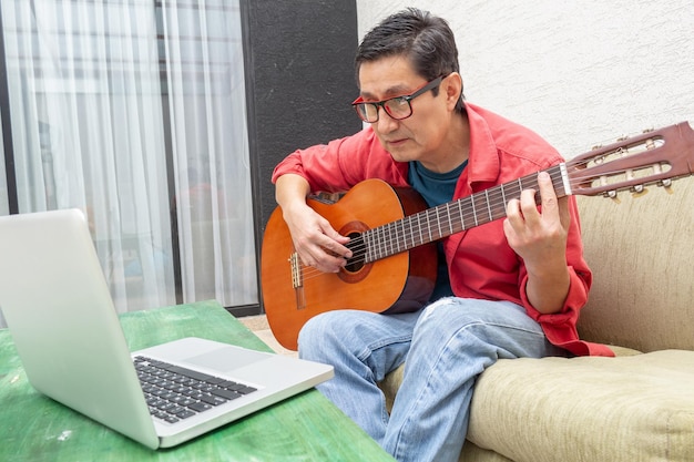 Homem olha atentamente para seu computador durante uma aula virtual de violão em casa