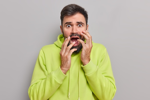 Foto homem olha intensamente para a câmera mantém a boca bem aberta sente-se alarmado com algo que tem grandes problemas observa algo assustador usando moletom verde posando na parede cinza