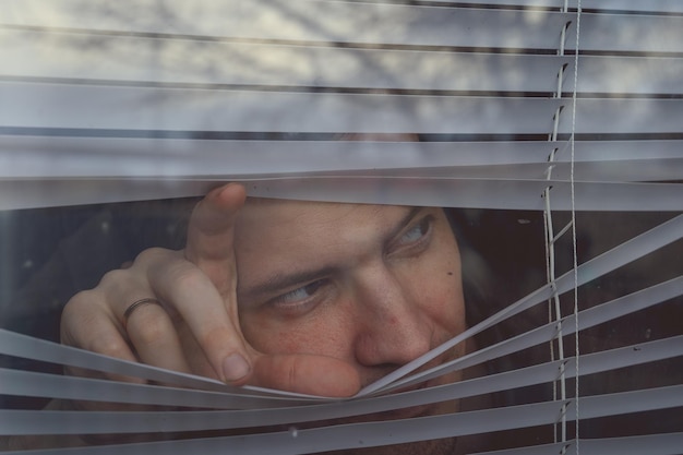 Homem olhando através das cortinas da janela Retrato de um jovem homem pensativo com olhos castanhos, observando através da persiana da janela