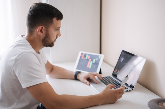 Homem olhando gráfico de finanças no laptop e comerciante do dia do tablet, investindo de monitores jovens em casa