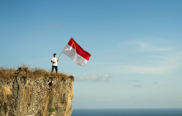 Foto homem orgulhoso da indonésia em um penhasco de praia erguendo bandeira vermelha e branca da indonésia