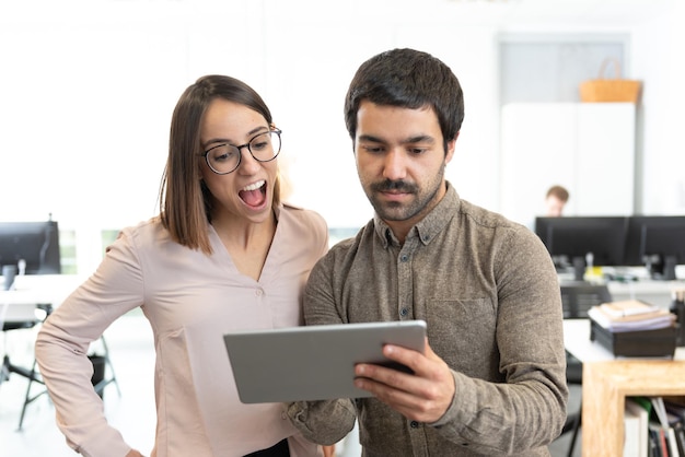 Homem orgulhoso hispânico mostrando algo para um colega de trabalho surpreso em seu tablet no escritório.