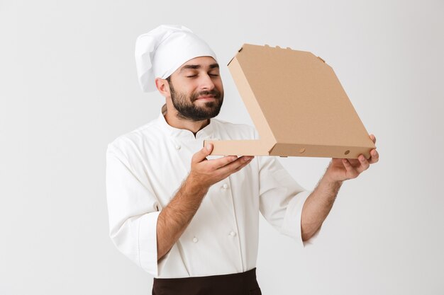 Homem padeiro feliz em uniforme de cozinheiro cheirando enquanto segura uma caixa de pizza isolada na parede branca