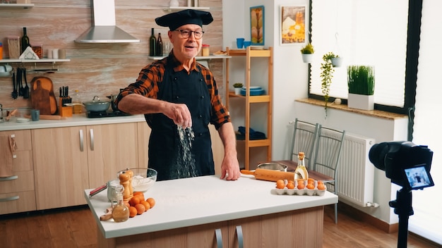 Homem padeiro gravando um videoblog de culinária enquanto prepara pizza em casa. Influenciador chef de blogueiro aposentado que usa a tecnologia da Internet para se comunicar, filmando blogs nas redes sociais com equipamento digital