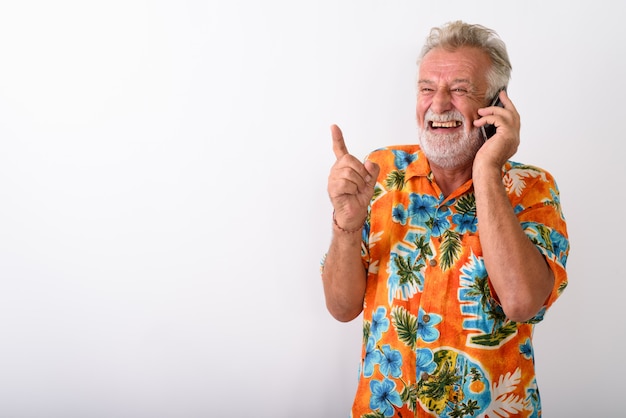 Foto homem pensativo e feliz turista barbudo sênior sorrindo e rindo enquanto fala no celular e aponta o dedo no branco