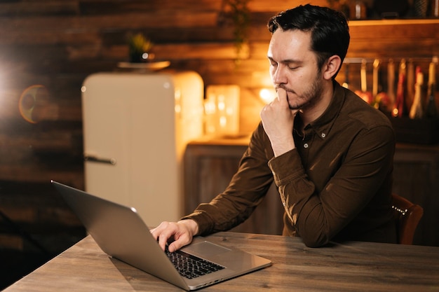 Homem pensativo em roupas casuais digitando no laptop enquanto está sentado à mesa