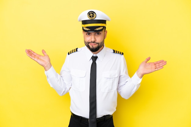 Homem piloto árabe de avião isolado em fundo amarelo, tendo dúvidas ao levantar as mãos