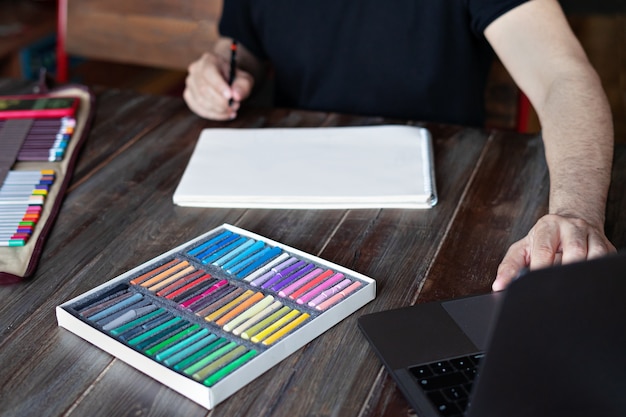 Homem pintando com lápis e giz de cera pastel no papel na frente do laptop