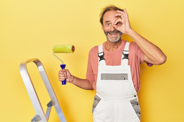 Foto homem pintando pano de fundo amarelo em estúdio animado mantendo o gesto ok no olho