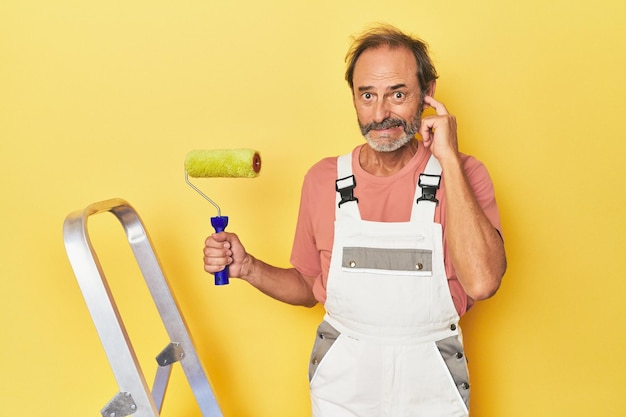 Homem pintando pano de fundo amarelo em estúdio cobrindo os ouvidos com as mãos