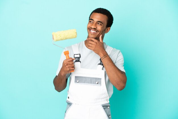 Homem pintor afro-americano sobre fundo azul isolado, olhando para o lado e sorrindo