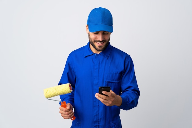 Homem pintor segurando um rolo de pintura isolado na parede branca, enviando uma mensagem com o celular