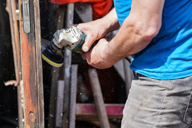 Foto homem polindo porta de madeira com moedor de ângulo antigo durante o dia ensolarado detalhe de close-up para mãos sem luvas poeira de madeira fina voando no ar